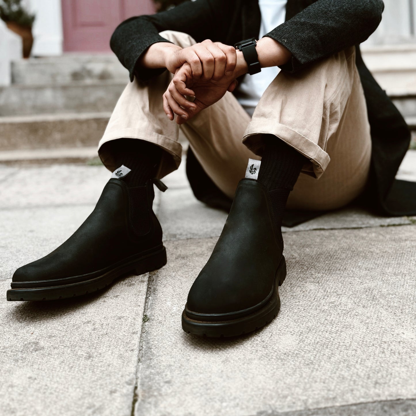 Man wearing black outdoor Chelsea boots, sitting in front of a colourful door in an urban setting.