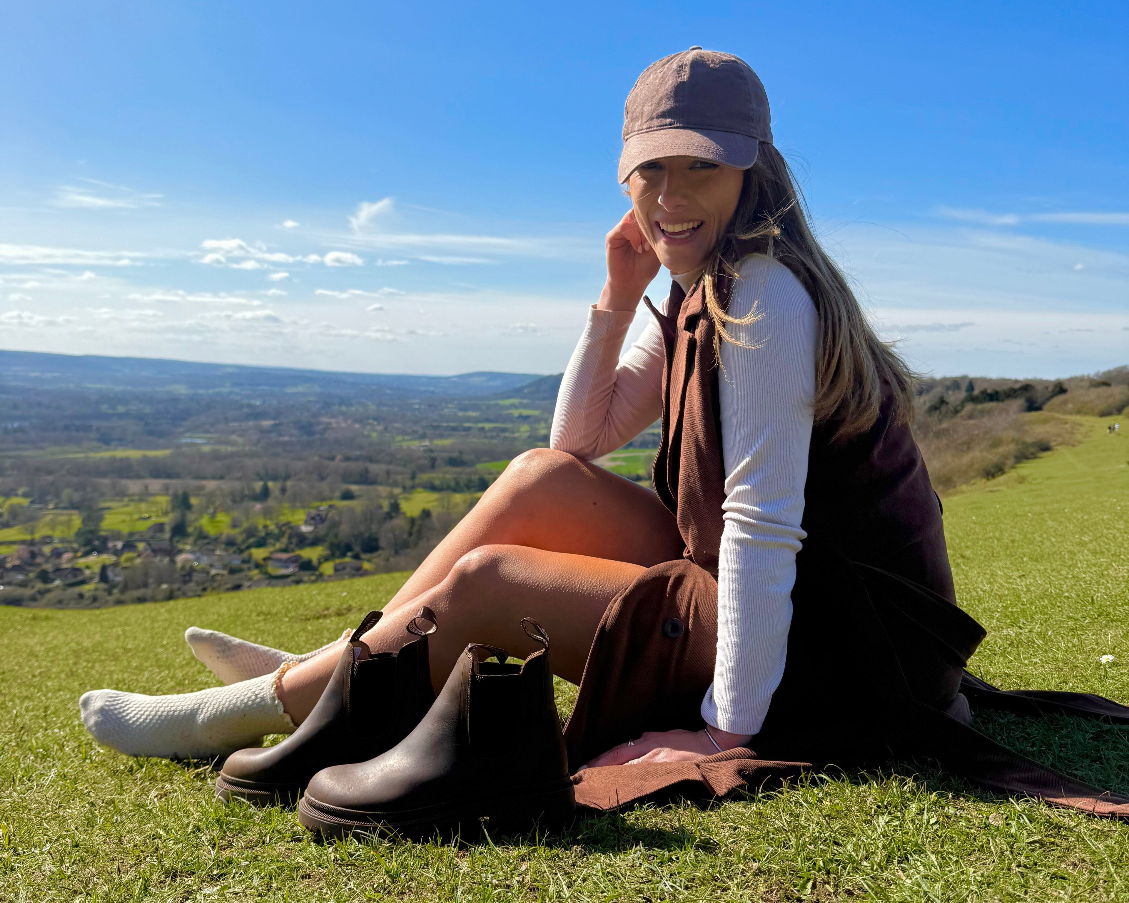 Woman sitting on a grassy hill with a scenic view, wearing a brown dress and brown chelsea boots next to her.
