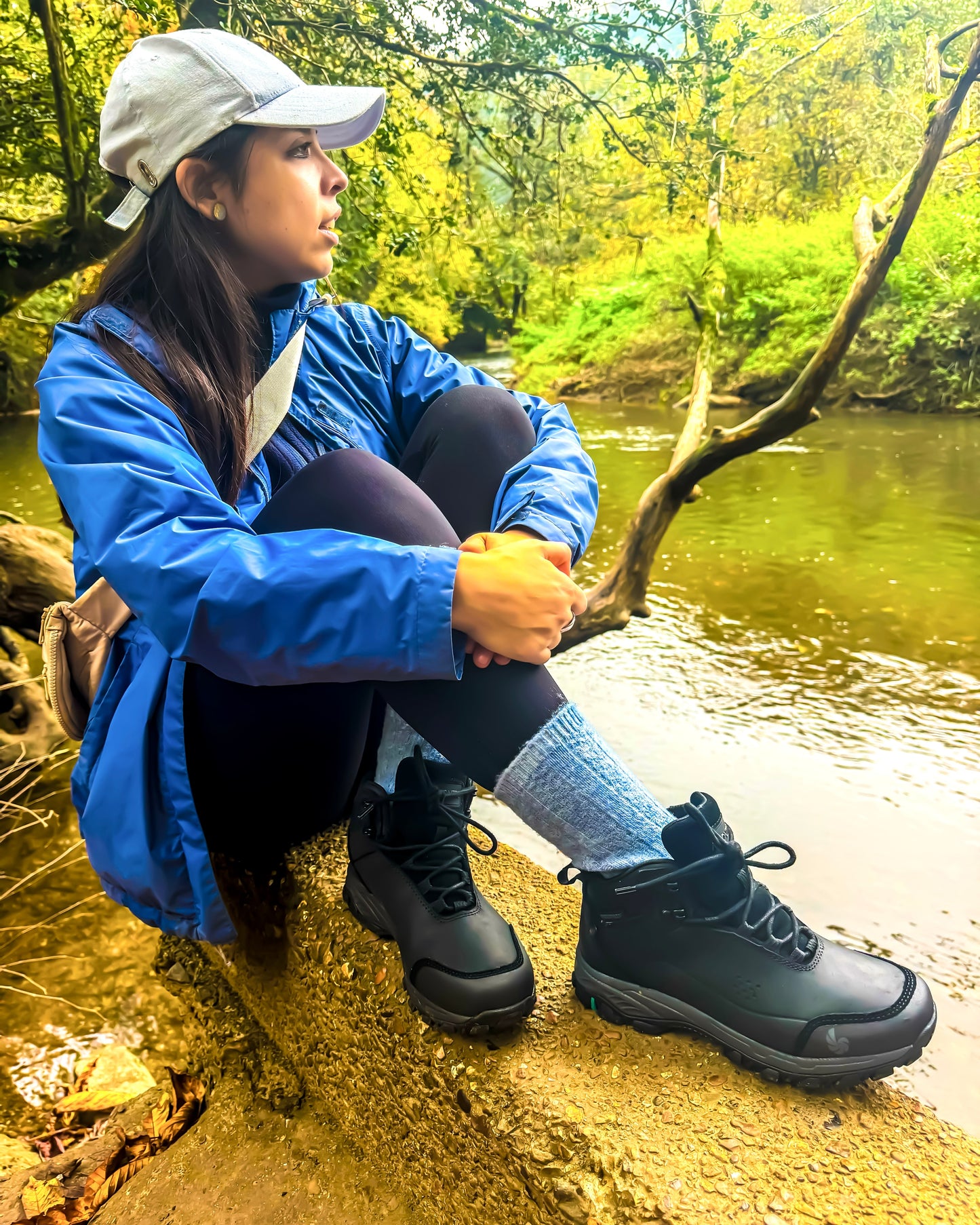 Woman sitting by a river wearing a blue jacket and black runswick hiking boots, surrounded by greenery.