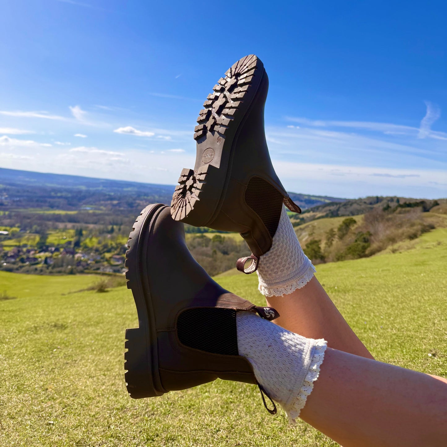 Woman wearing brown chelsea boots and white socks with a scenic view of green fields and blue sky in the background.