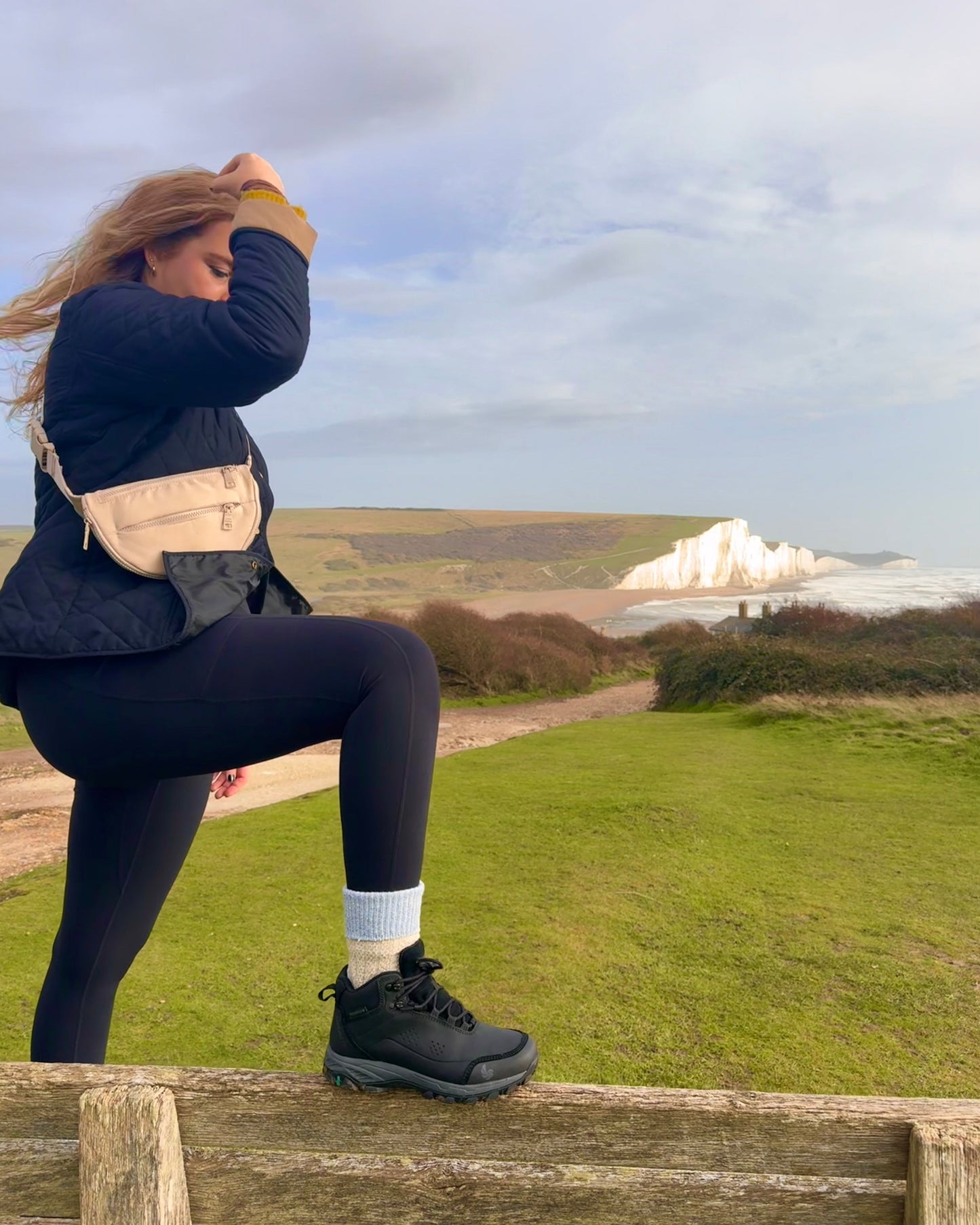 Woman standing on a wooden bench wearing black hiking boots with a scenic view of a coastal landscape in the background