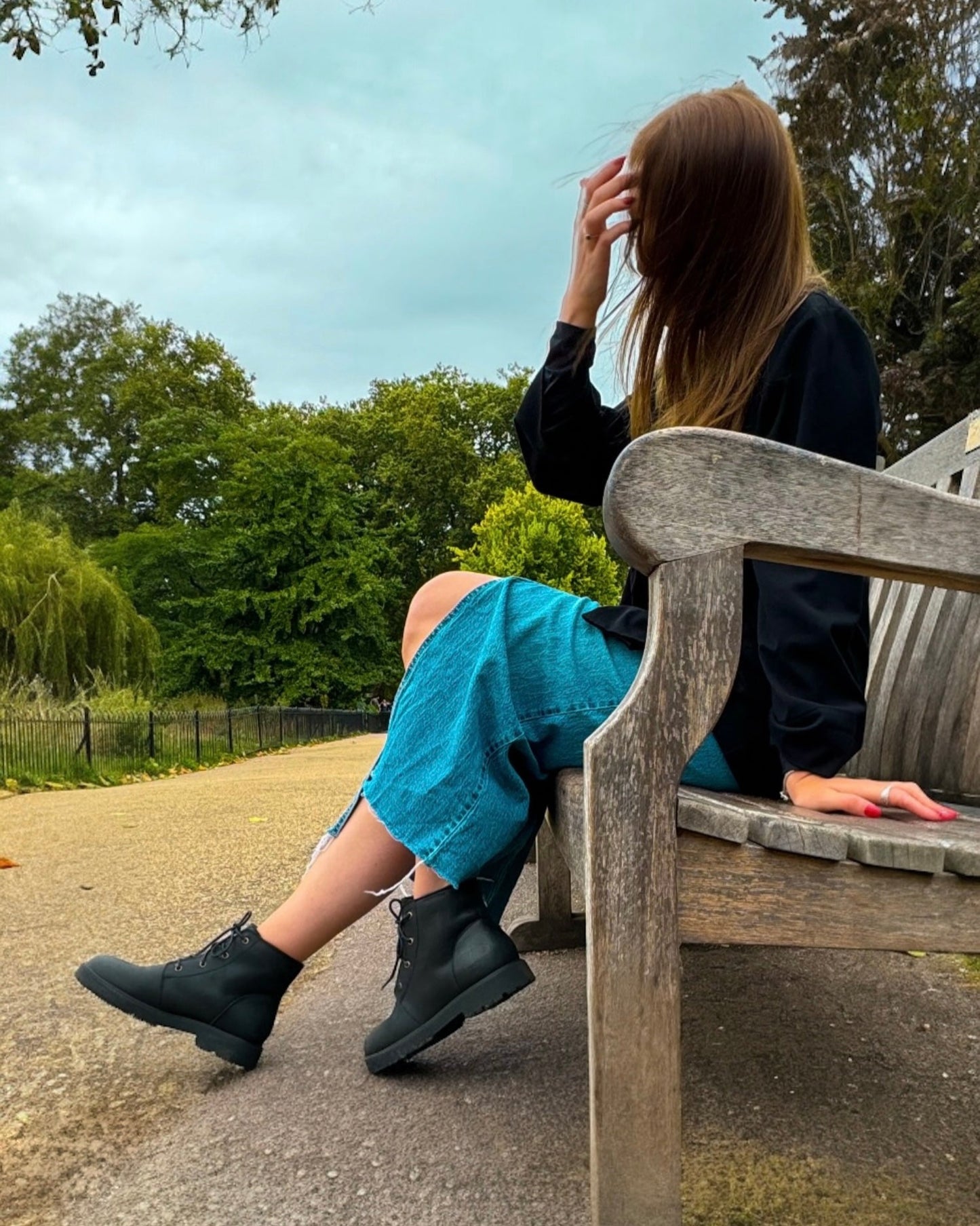 Woman sitting on a bench in a park with trees and a cloudy sky in the background showcasing black all-terrain boots.