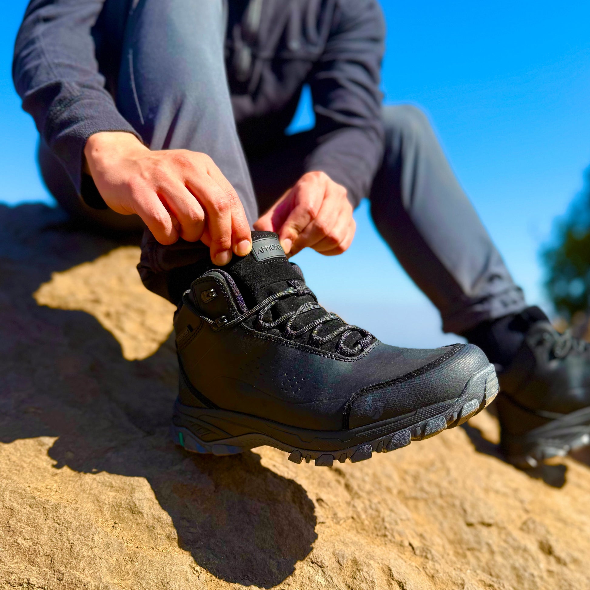 Person tying a black hiking boot on a rocky surface with a clear blue sky.