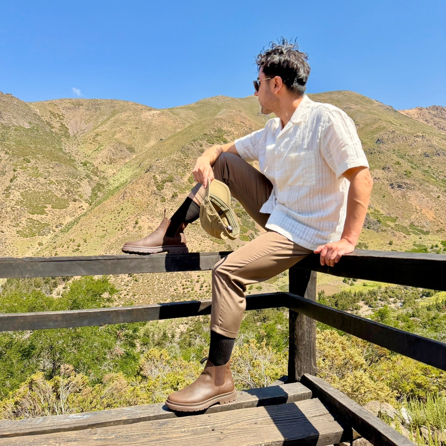 Person sitting on a wooden railing overlooking a mountainous  landscape, wearing brown chelsea boots