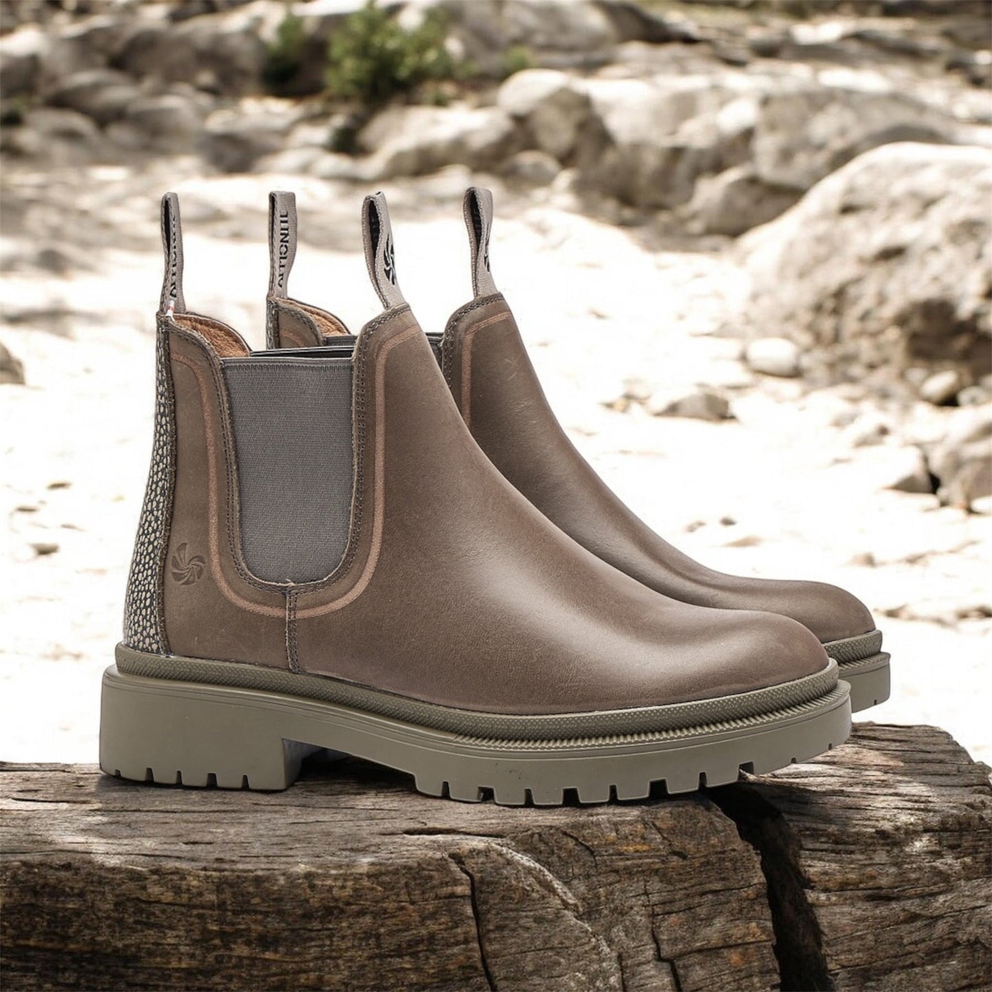 Pair of grey Chelsea boots on a wooden log with a natural stone background