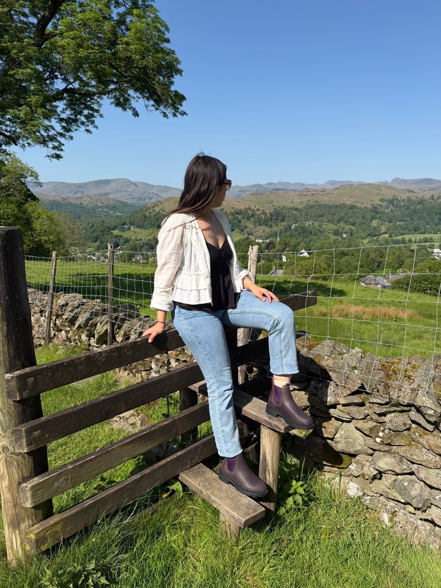 Person sitting on a wooden fence in purple chelsea boots overlooking a scenic landscape with mountains and green fields.